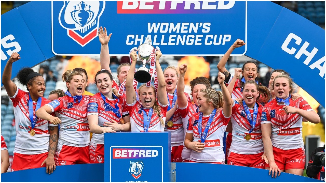St Helens' women's team hold the Challenge Cup trophy aloft, celebrating their victory in white shirts with a red V.