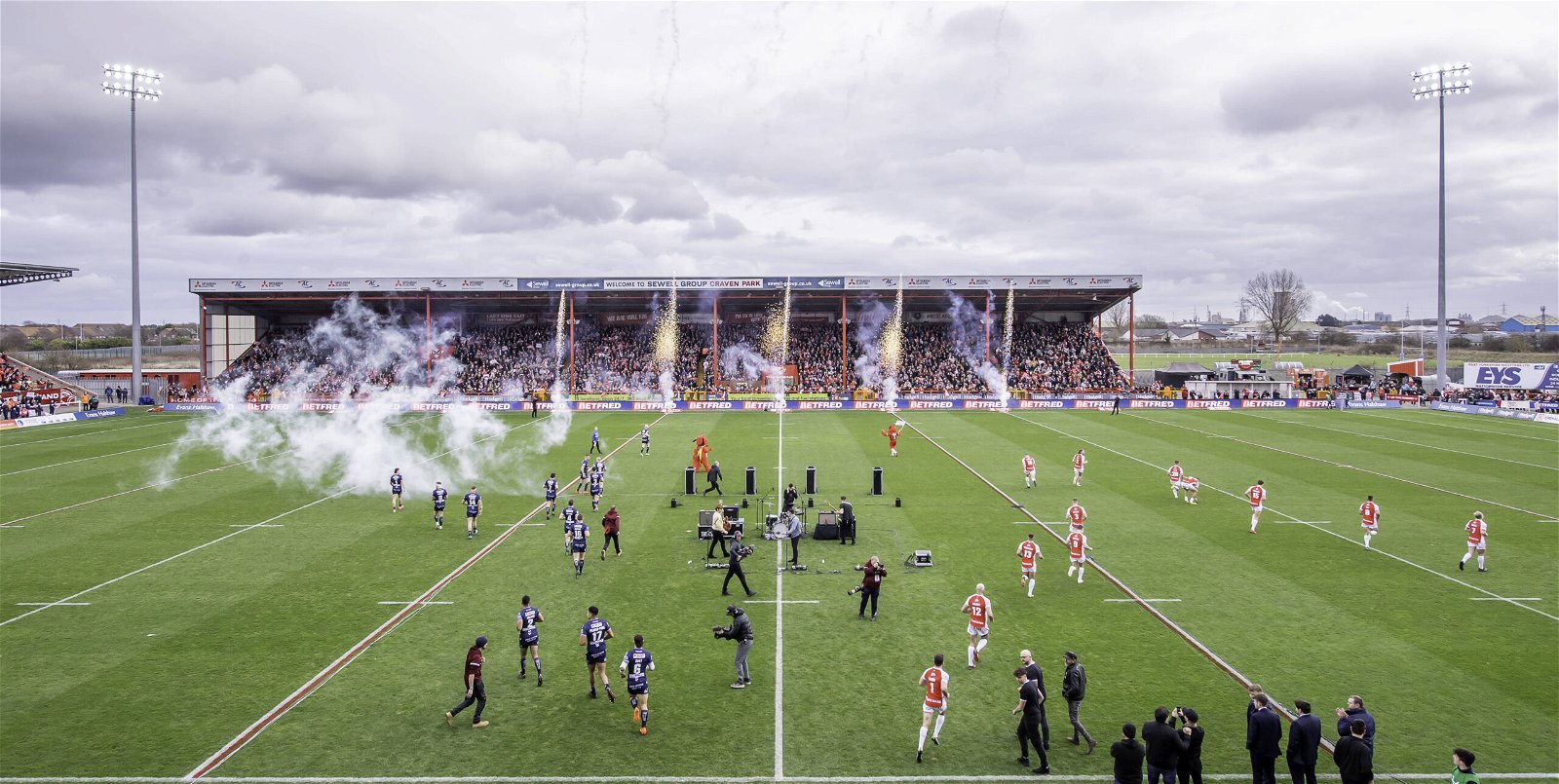 A wide shot, as Hull KR and Wigan come out to the field of play with pyrotechnics at Craven Park