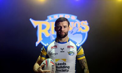 Andy Ackers in studio light models the new Leeds Rhinos kit at the Super LEague launch day. It's white (yeah) with blue collar and blue sleeves. The sleeves have a yellow stripe on, too. The AMT logo, Berry's cursive logo and OXEN can be seen. Leeds Building Society returns as main sponsor.
