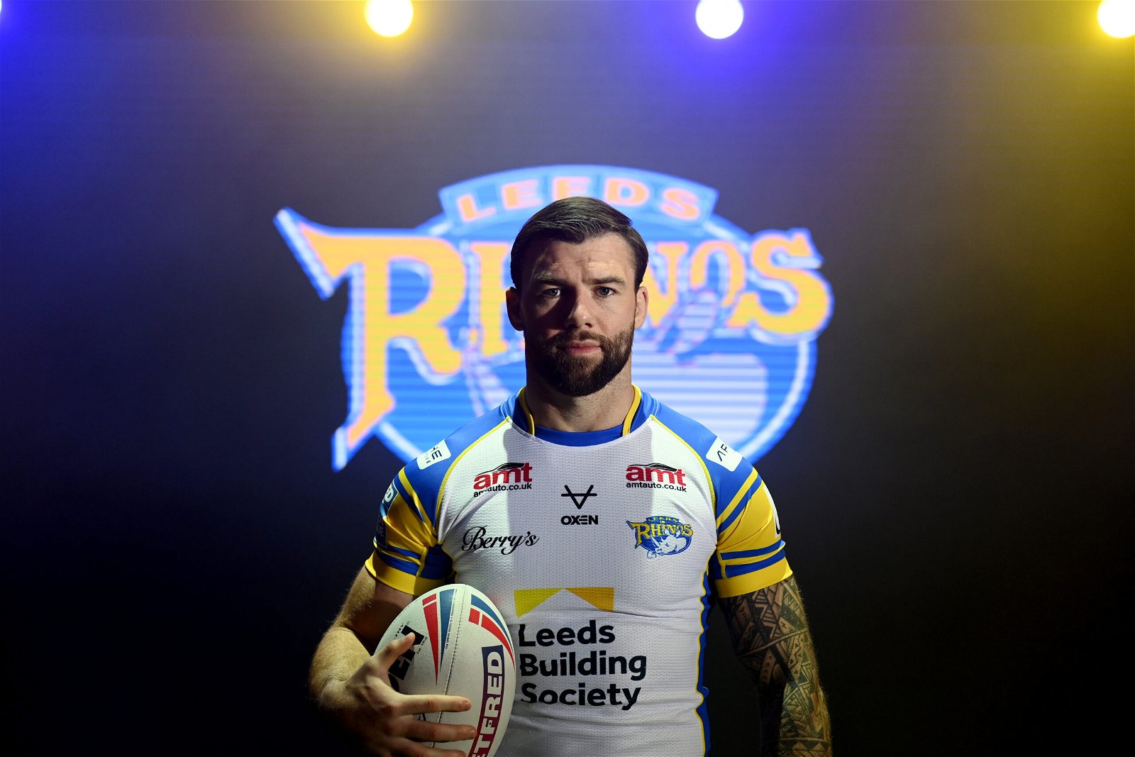 Andy Ackers in studio light models the new Leeds Rhinos kit at the Super LEague launch day.It's white (yeah) with blue collar and blue sleeves. The sleeves have a yellow stripe on, too. The AMT logo, Berry's cursive logo and OXEN can be seen. Leeds Building Society returns as main sponsor.