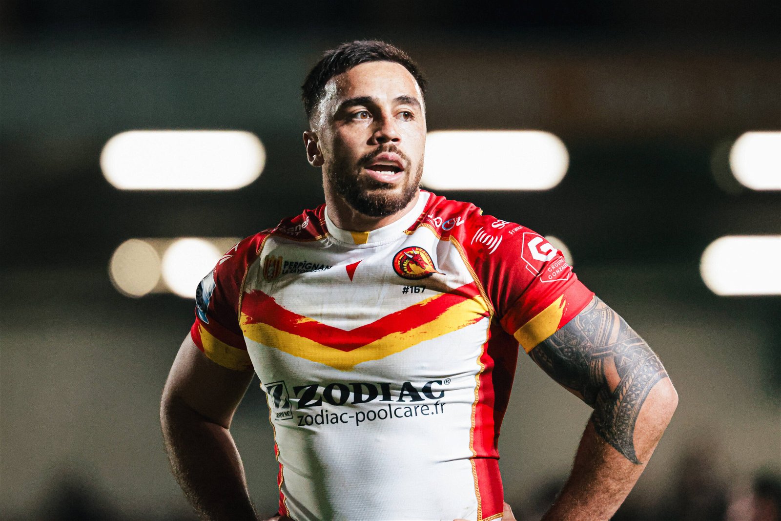 Matt Ikuvalu wearing a Catalans shirt - it's white, with a thin red and yellow chevronbelow the badge, Macron logo and 'Perpignan La Rayonnante' logos.The Zodiac logo below is black. The shoulders and sleeves are red, with a little bit of yellow in a brush-stroke style kind of thing going on.