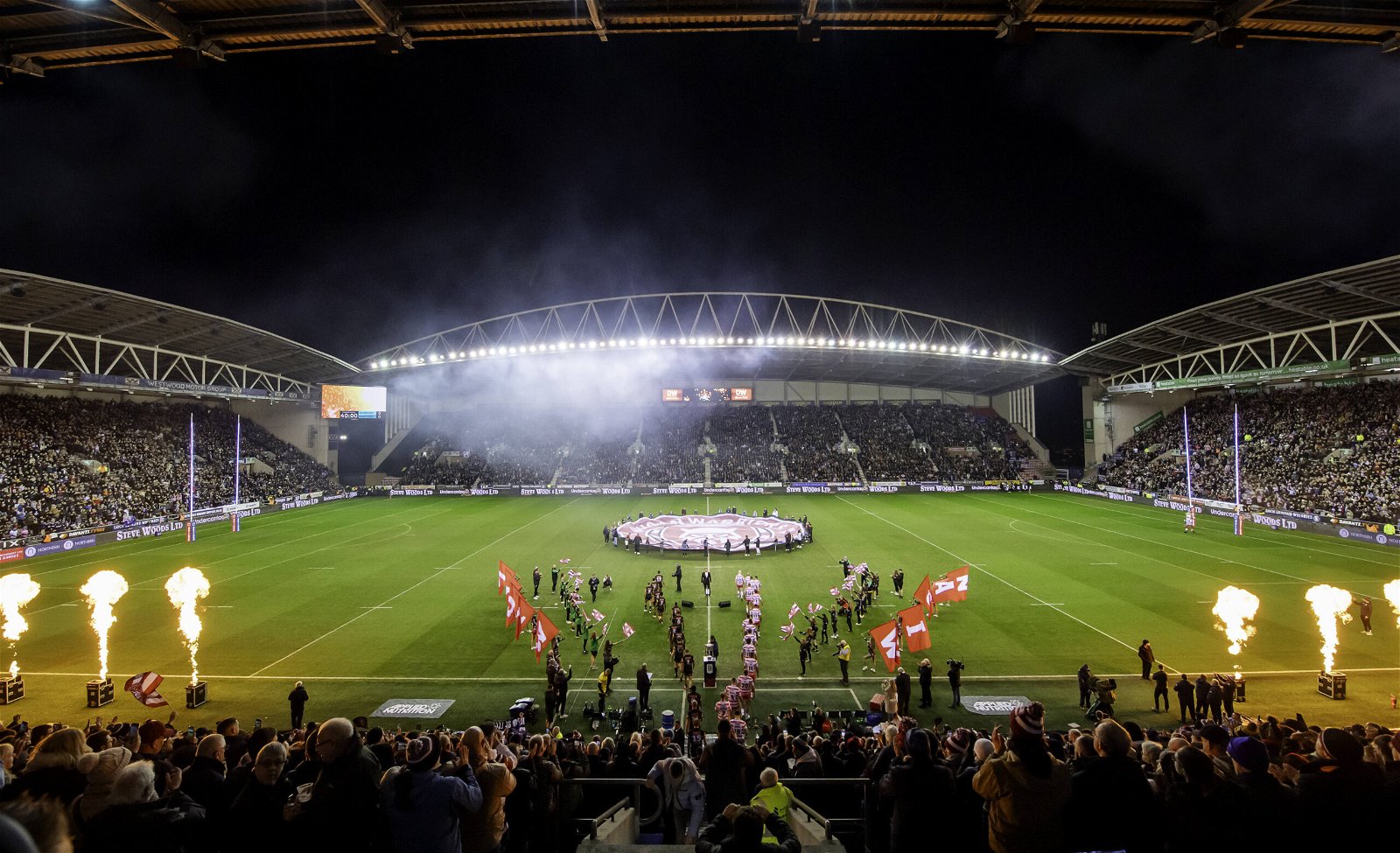 A view of the stand opposite the main stand at DW Stadium before the World Club Challenge 2024. Players are walking out, a fire display can be seen, and the stands are full.