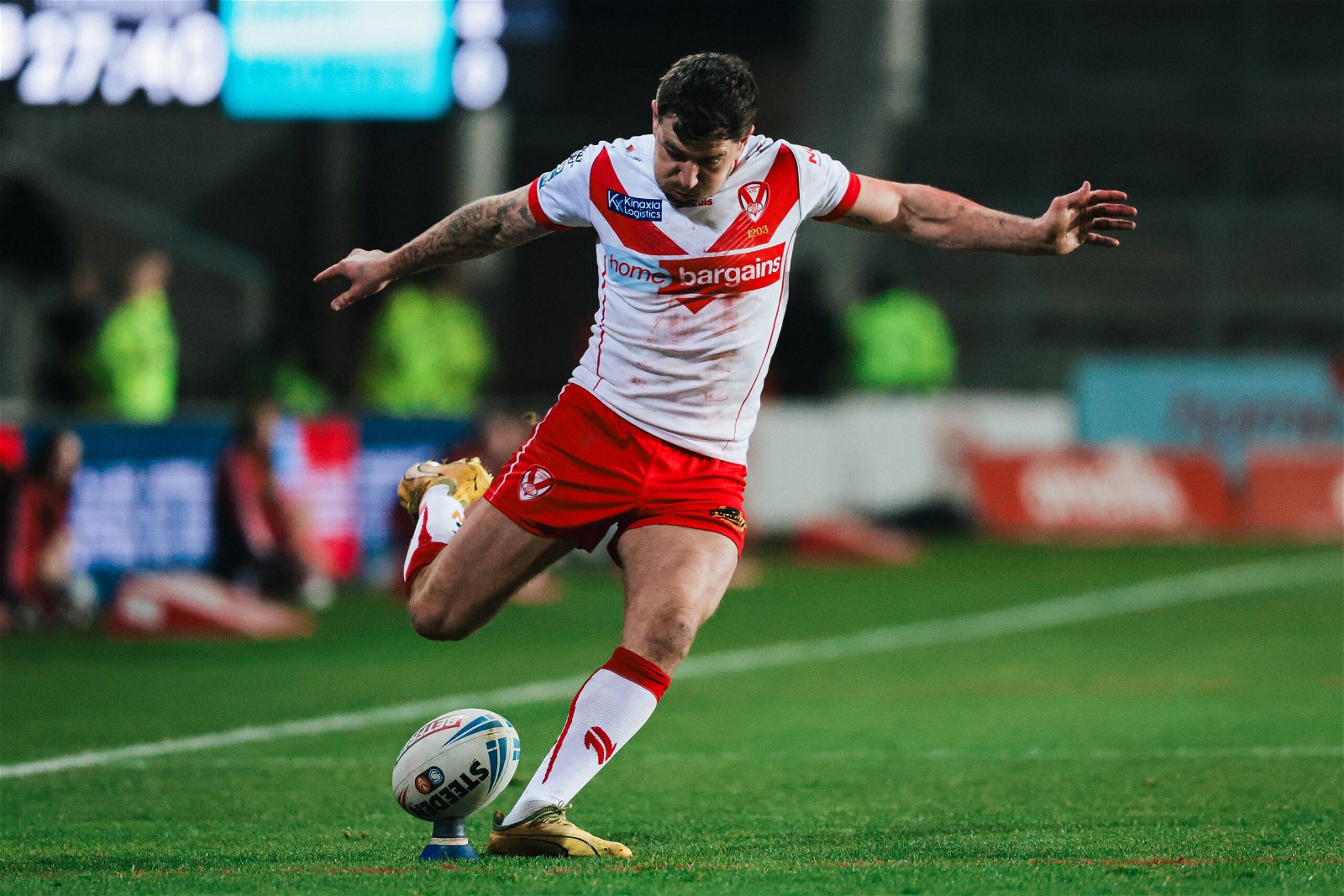 Mark Percival is wearing the St Helens shirt, kicking for goal. Of course, it's white with a red V, and there's a thin red line down the sides. The collar and sleeve cuffs are red. The Home Bargains logo on it is huge.