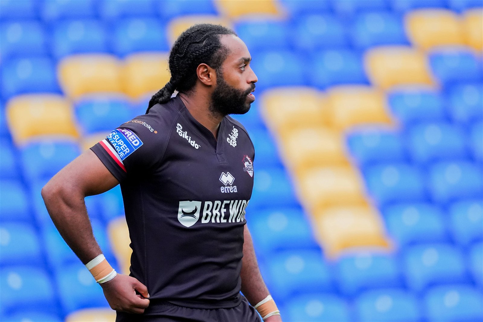 London Broncos' Iliess Macani looks to the side during Super League action.His shirt is a plain black one, with the club crest, the Brewdog logo as main sponsor, and barely legible sponsors by the collar.