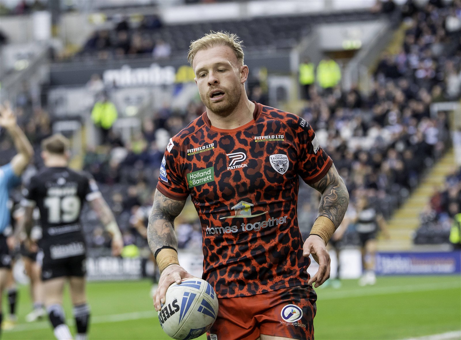 Leigh Leopards' Oli Holmes holds the ball having just scored a try.The shirt is red with black leopard print. Holland and Barrett, Fitfield Ltd and Homes Together logos can be seen (just about)!