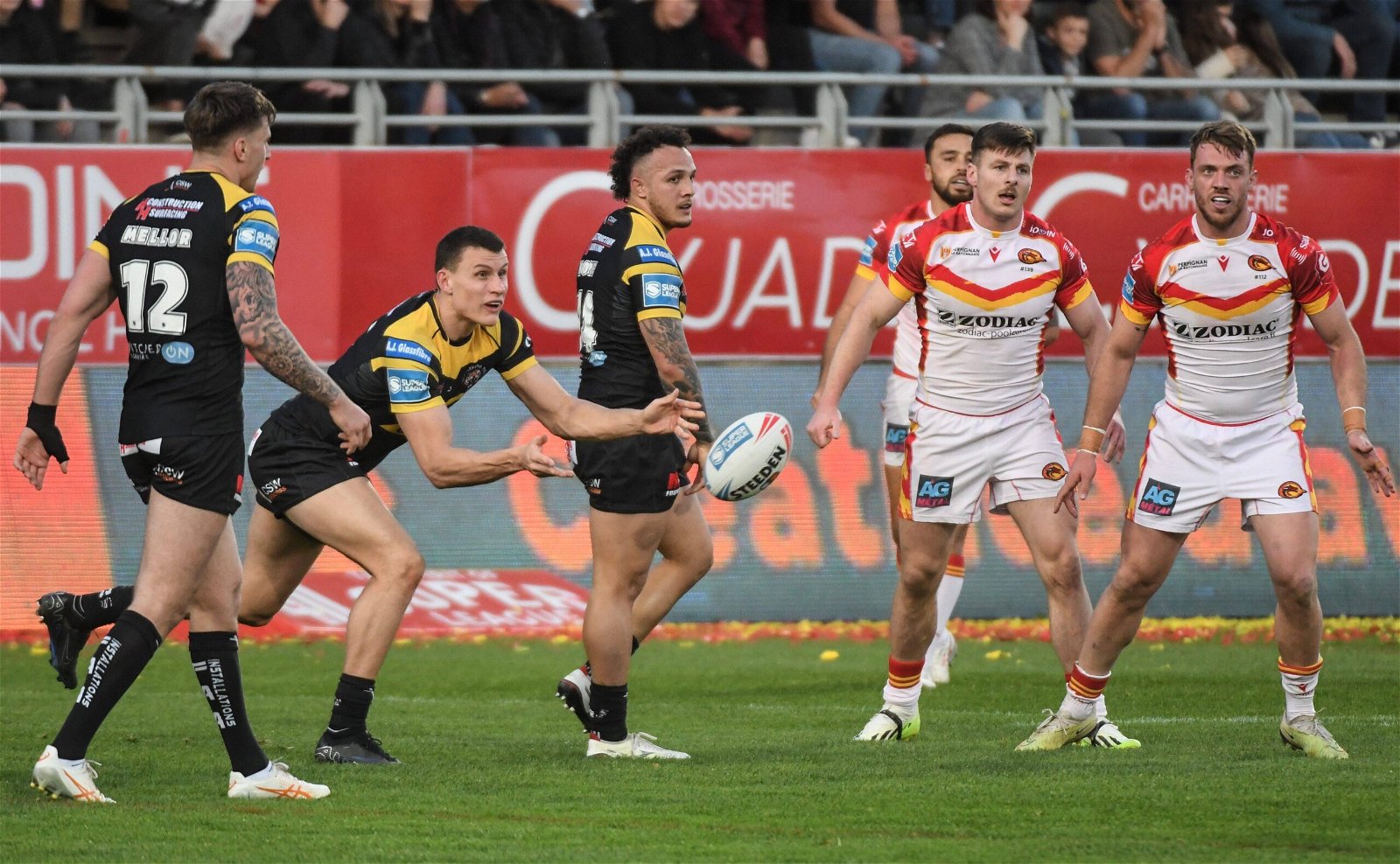 Innes Senior passes the ball for Castleford Tigers, in their black and amber kit, in Super League action against Catalans Dragons, who wear white with red and yellow detailing.